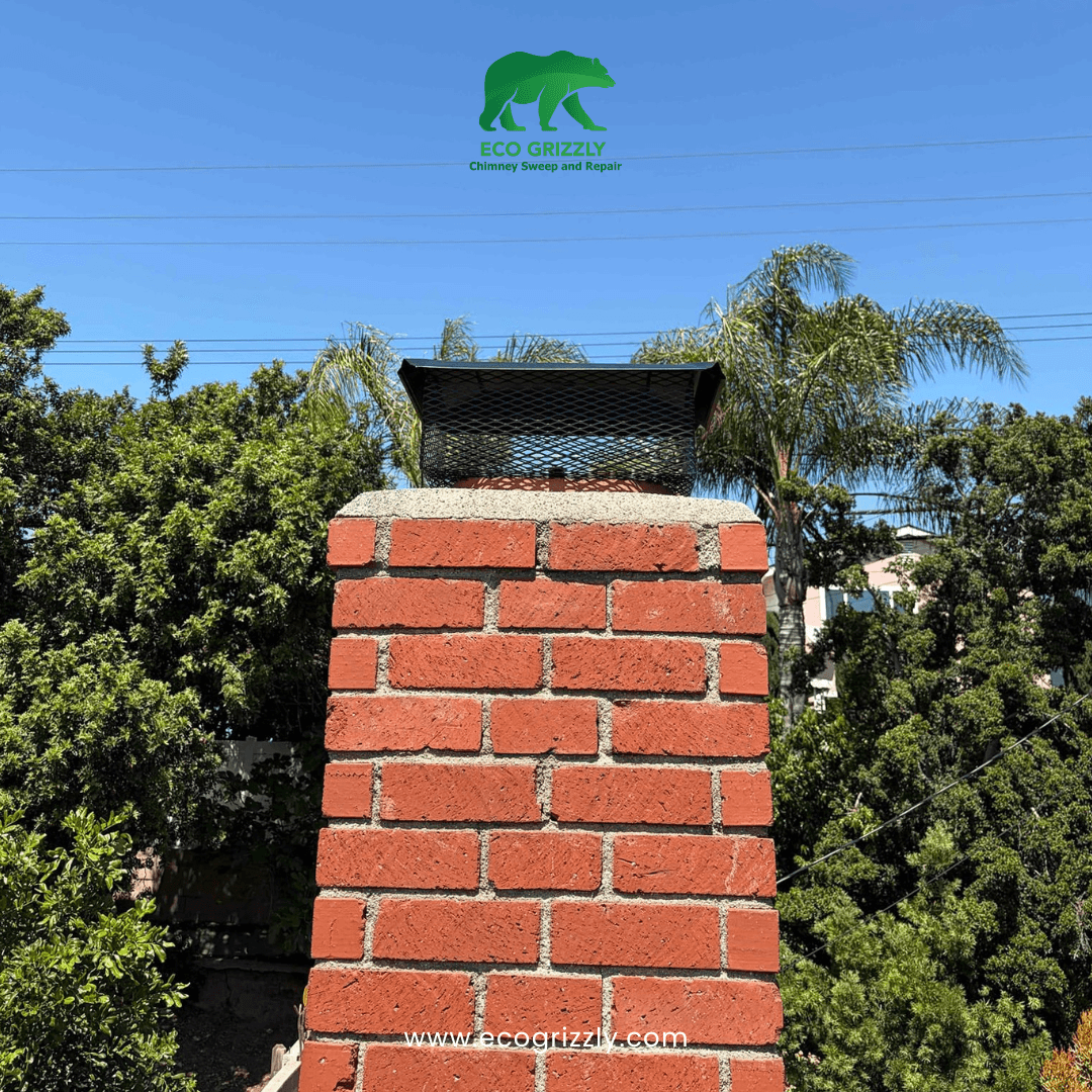 Red brick chimney with black mesh chimney cap installed on top surrounded by green trees