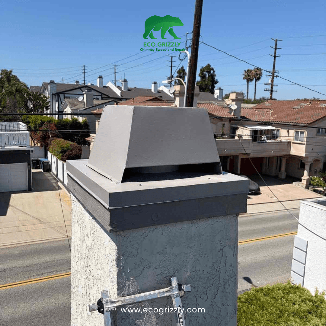 Gray angular chimney cap installed on a stucco chimney with ladder leaning against it