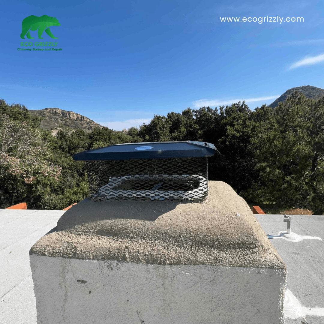 Black metal chimney cap on a stucco chimney with rolling hills and blue sky behind