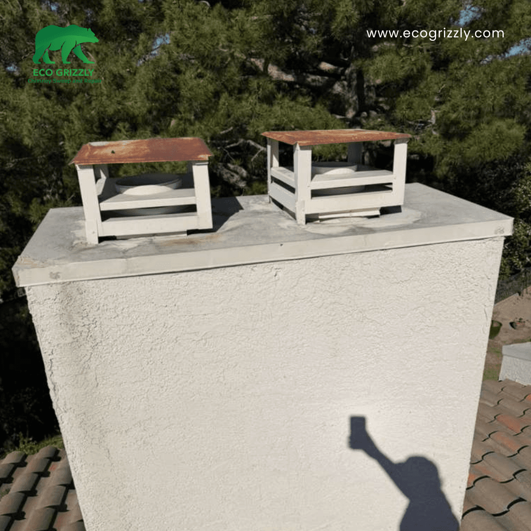 Two white chimney caps with rusty tops on a stucco chimney before replacement