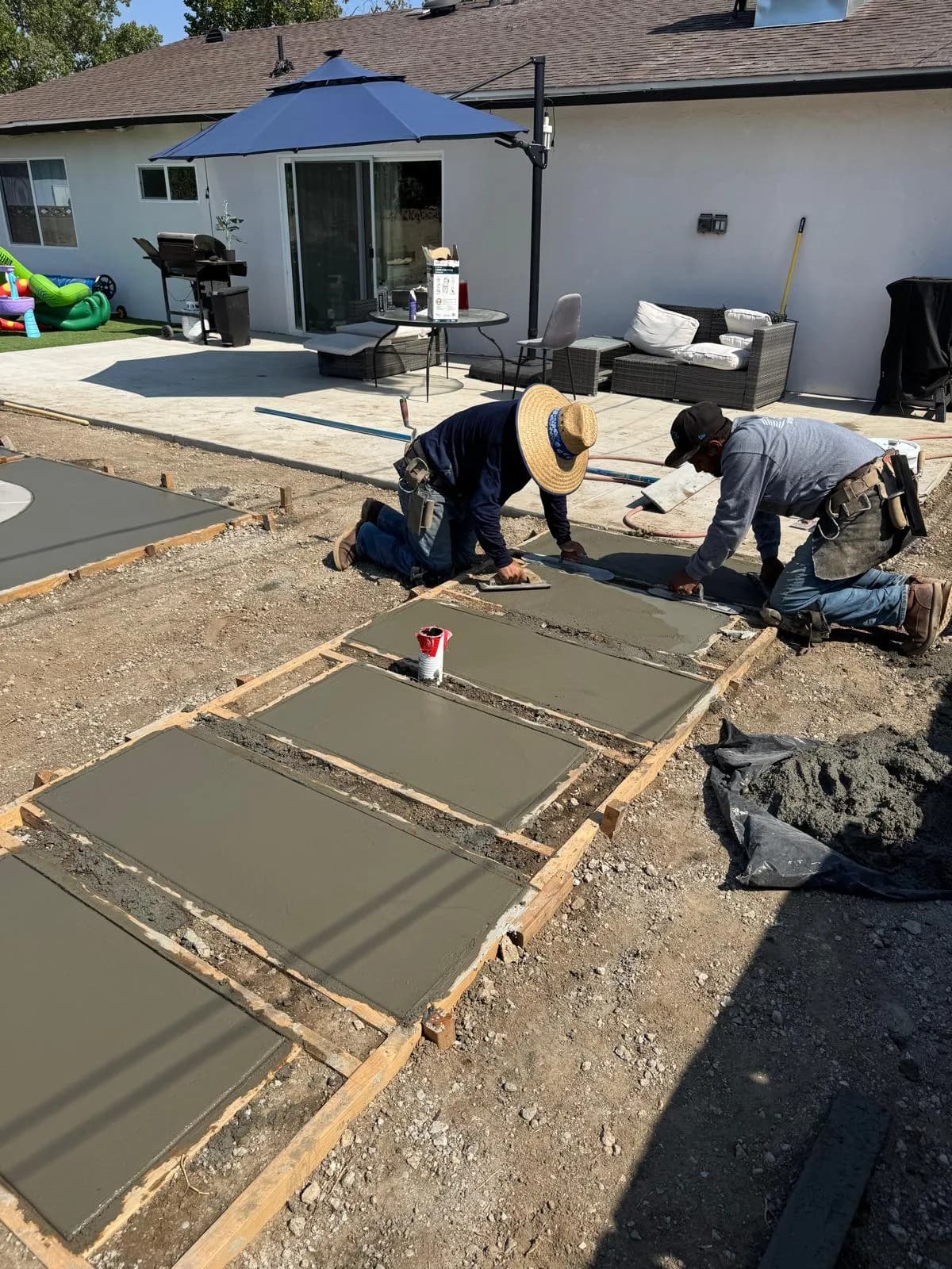 Two workers smooth wet concrete in framed sections along a backyard patio beside a house