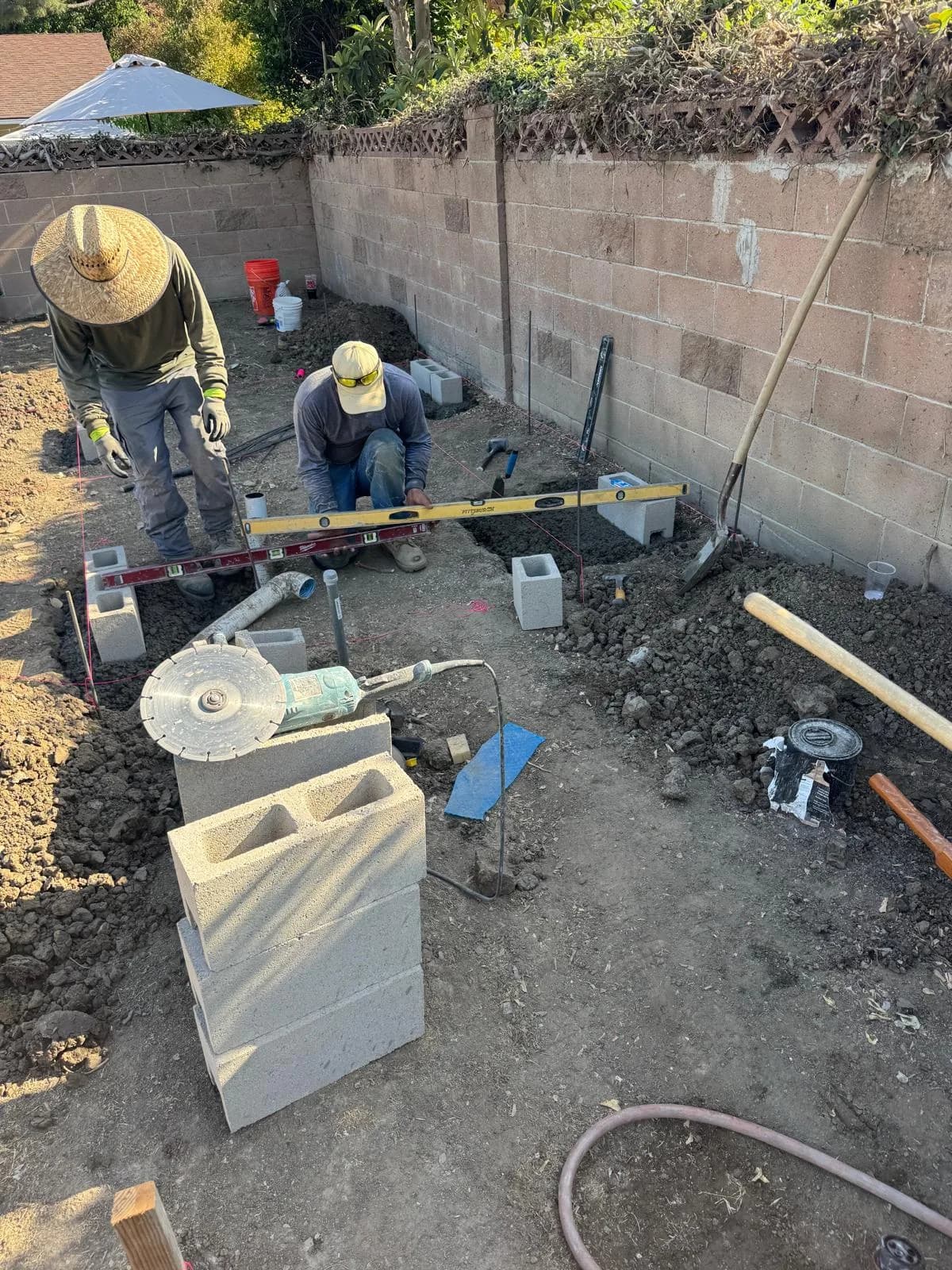 Two workers level cinder blocks in a dirt yard with tools and string lines for outdoor fireplace construction