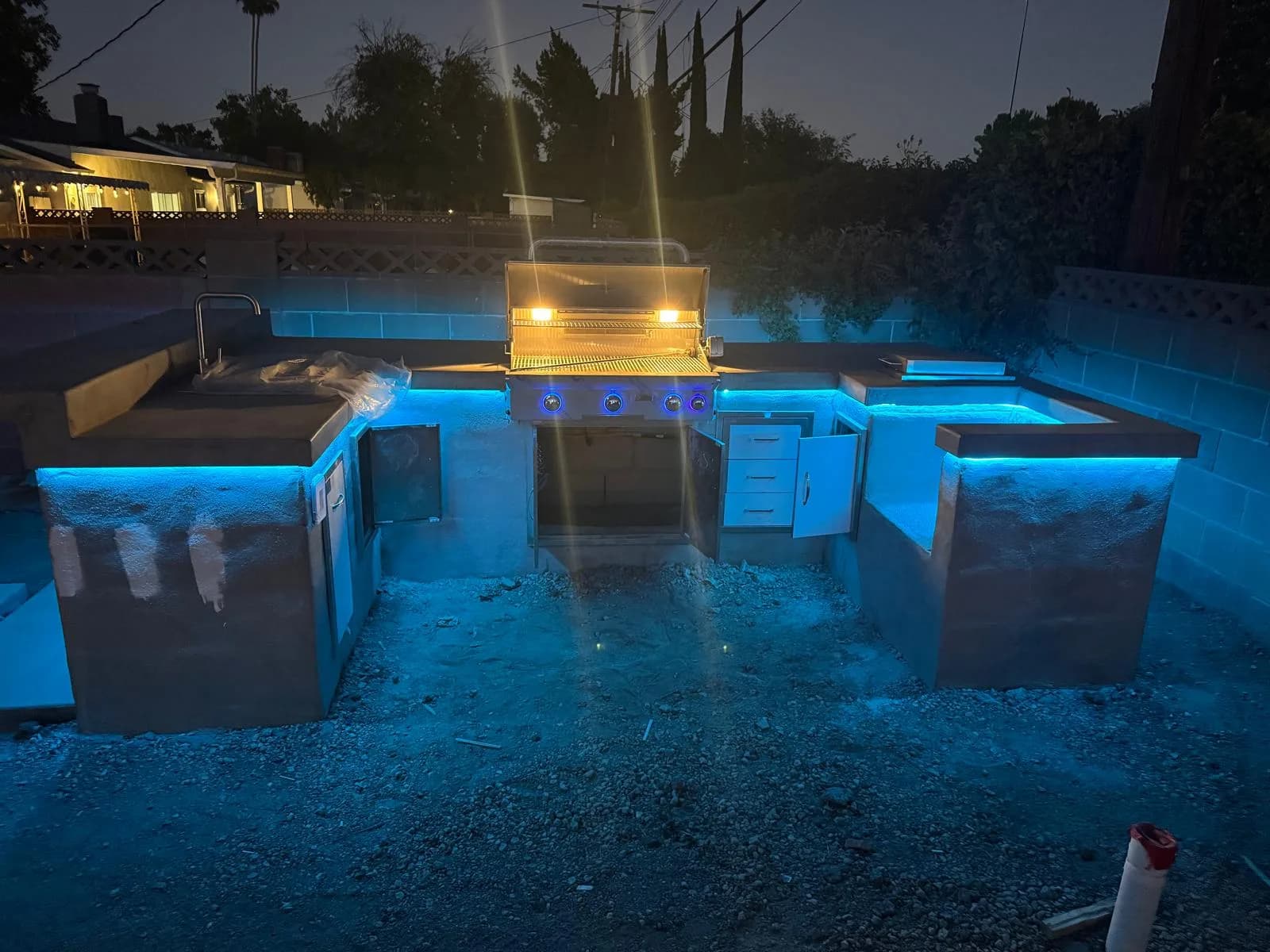 Outdoor kitchen under construction at night with lit grill, blue accent lighting, and unfinished ground