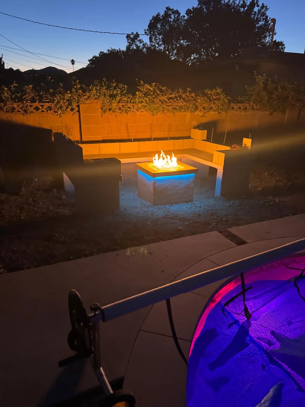 Finished backyard fire pit with built in bench seating glowing at dusk beside a lit pool edge