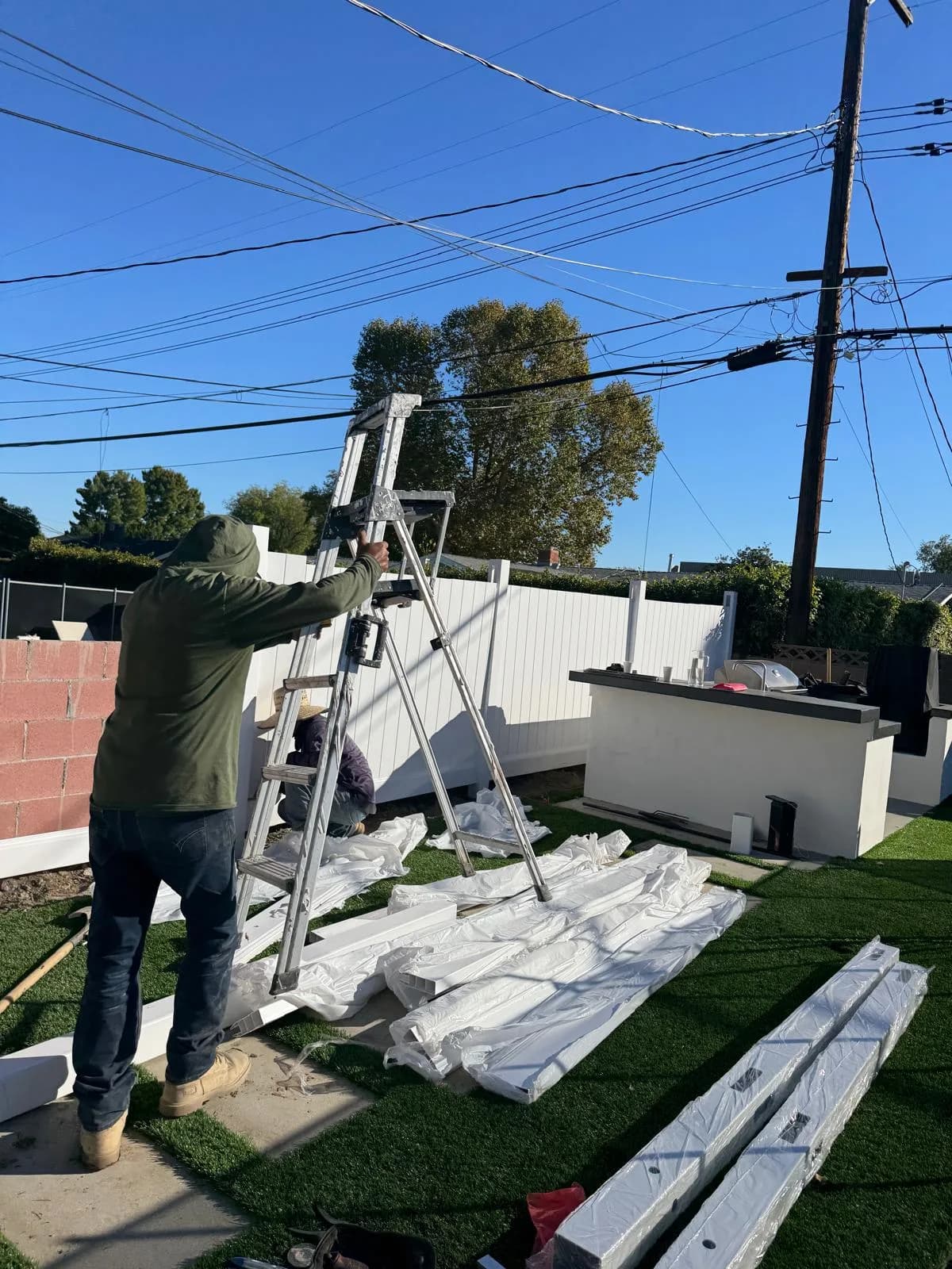 Worker sets up a ladder beside wrapped trim pieces in a backyard during outdoor remodel construction
