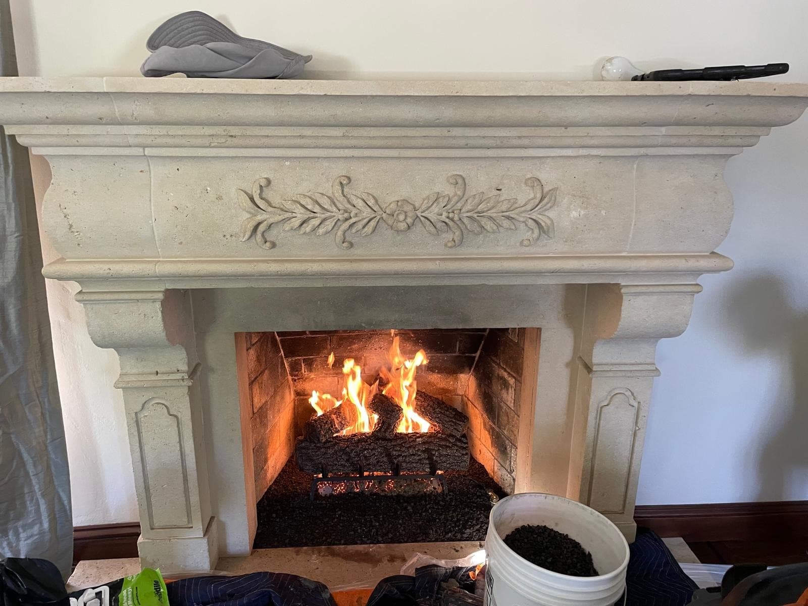 New gas log set burning in a firebox under a carved cast stone mantel with a bucket of lava rock nearby