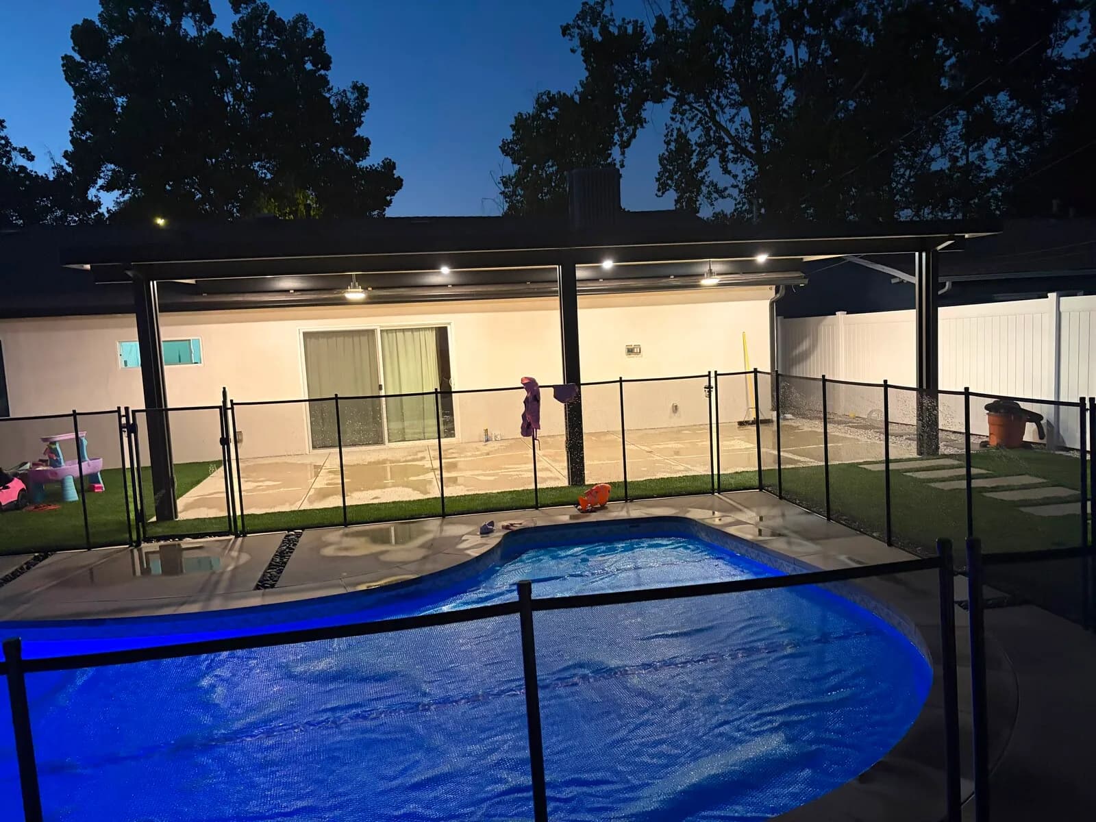 Backyard at dusk with a lit aluminum patio cover, covered pool and black mesh safety fence