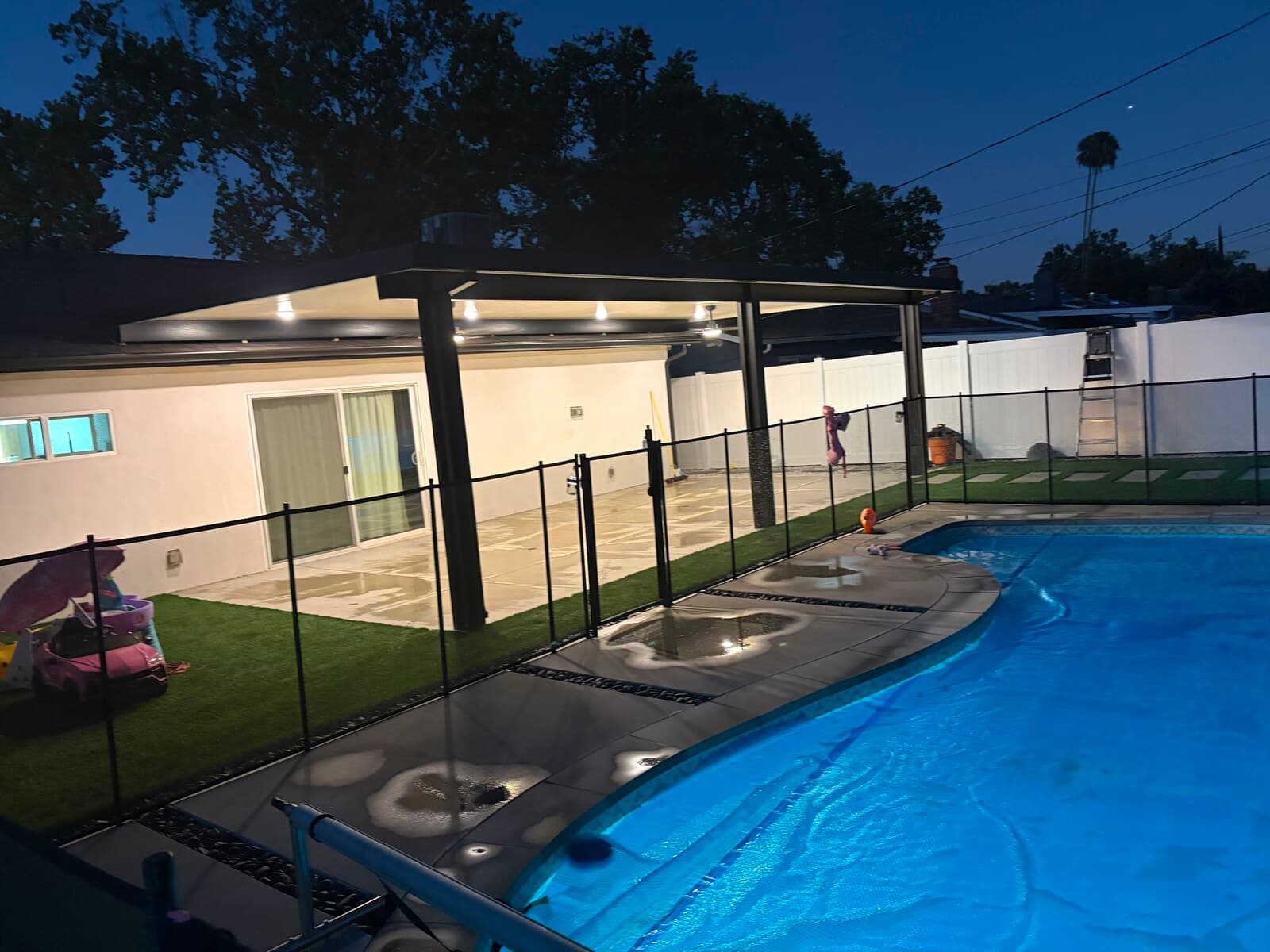 Angled night view of the lit patio cover, artificial turf and black safety fence along the glowing pool