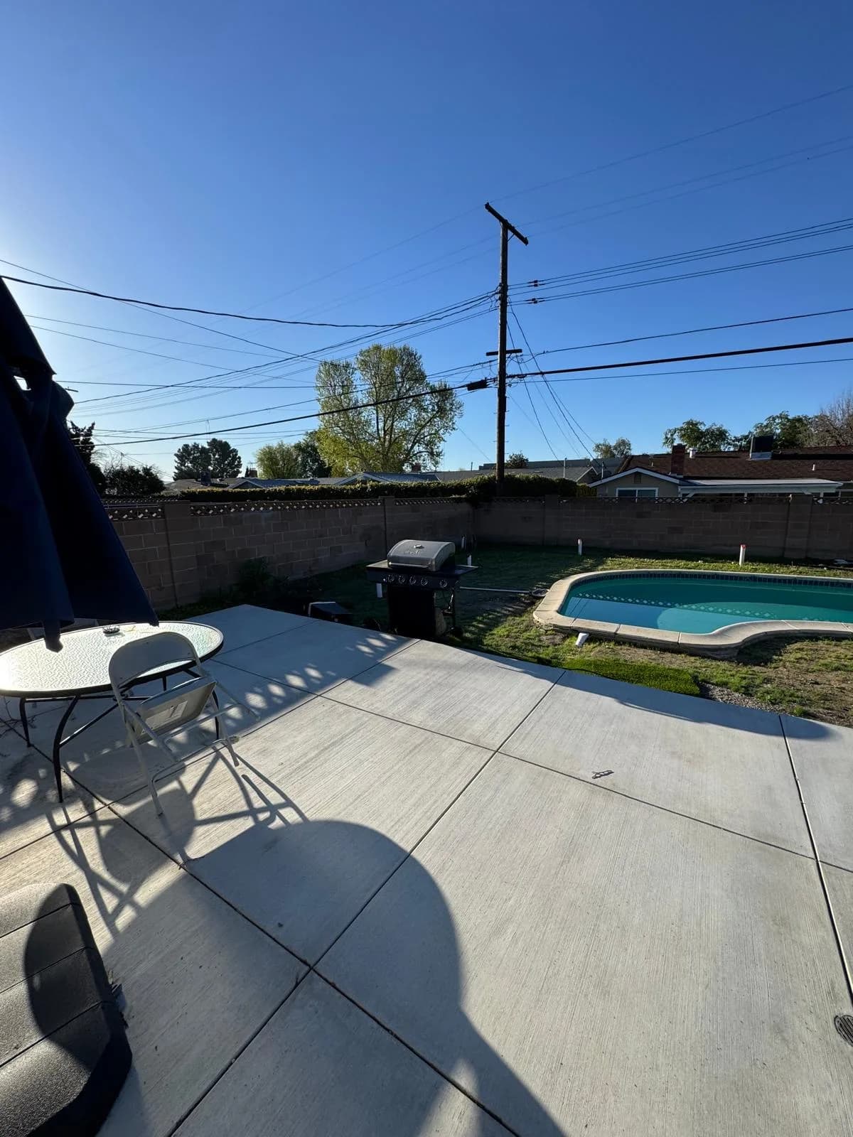 Backyard patio with a small swimming pool, grill, table, and chair beside a block wall under a clear sky