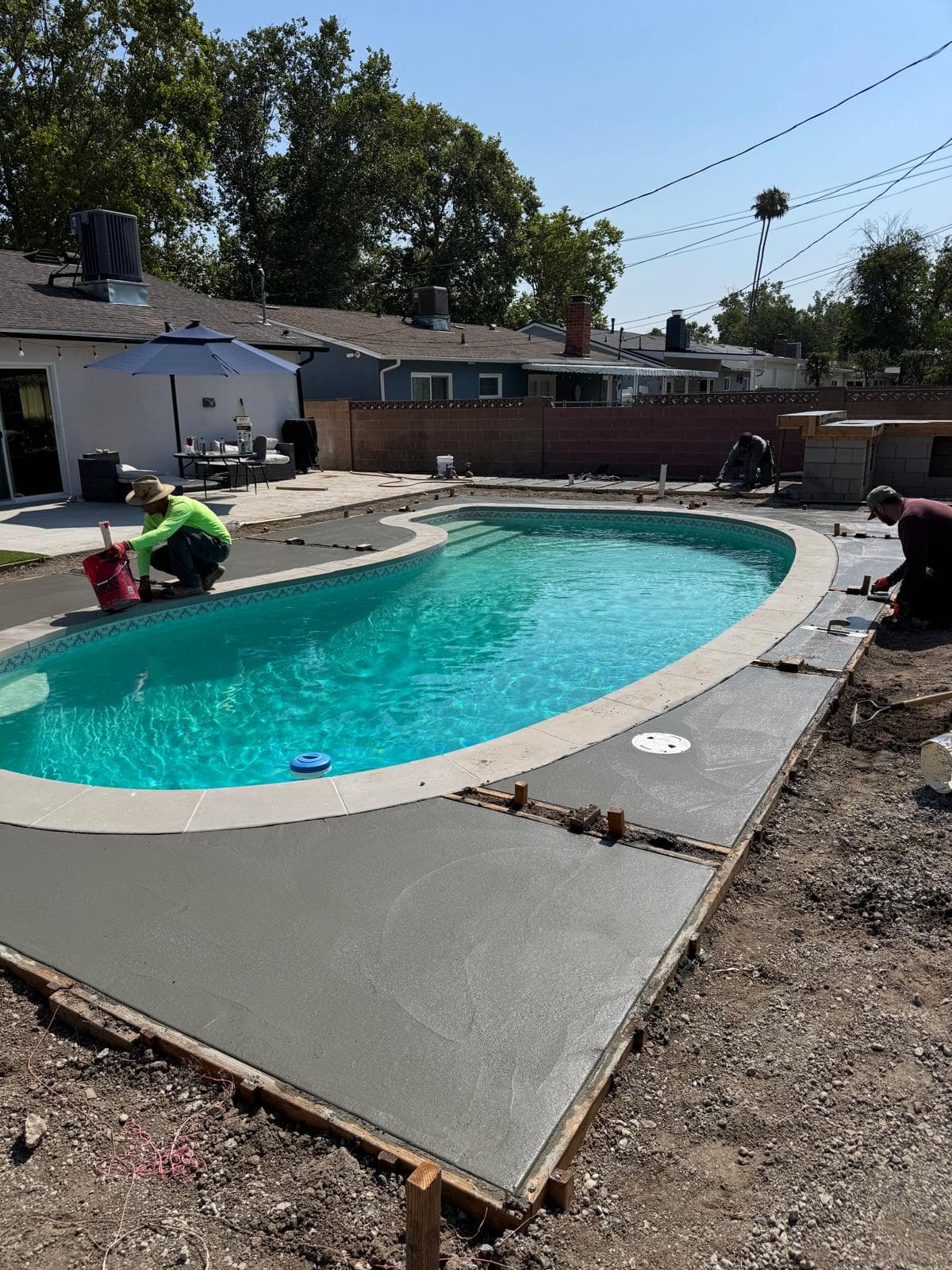 Workers pour and smooth new concrete decking around a residential swimming pool during remodel