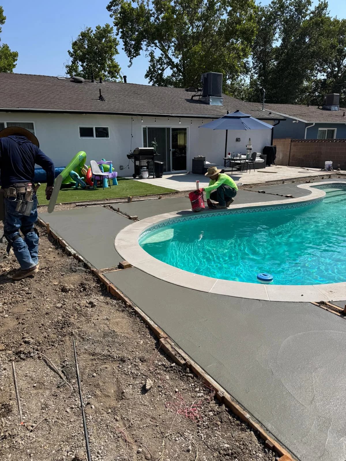 Workers pour and smooth new concrete decking around a residential swimming pool during remodel