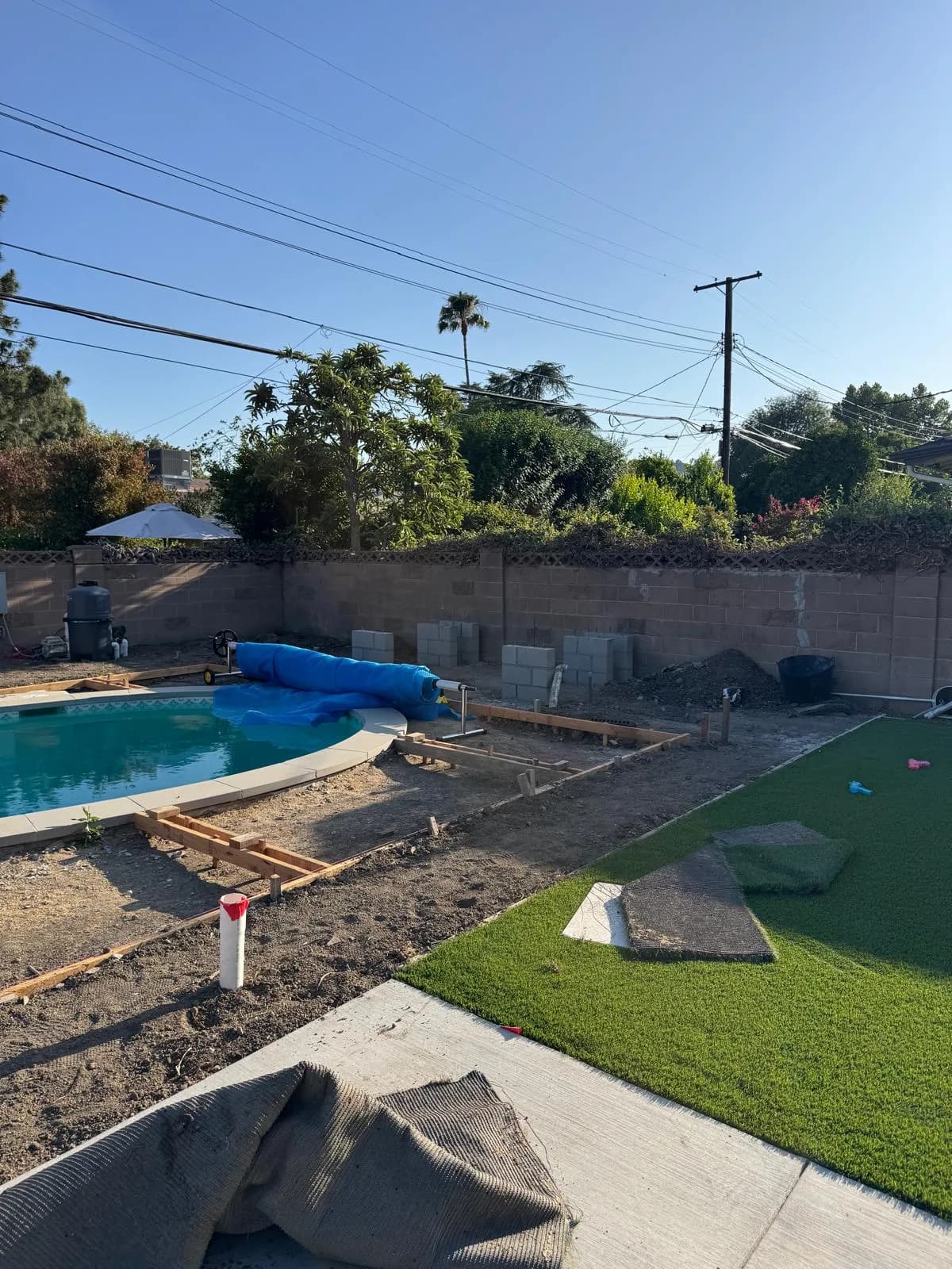 Pool remodel in progress with new deck forms, exposed soil, and rolled cover beside the water