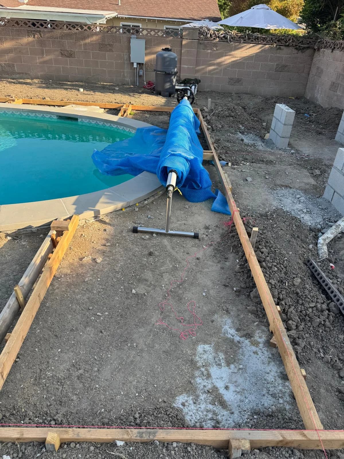 Wood forms outline a new pool deck beside the pool, with dirt grading and a rolled blue pool cover nearby