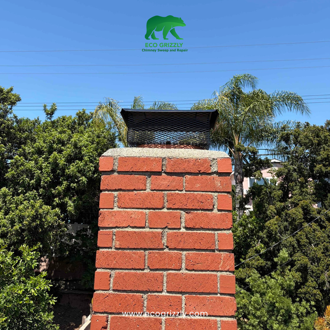 Red brick chimney with black mesh chimney cap installed on top surrounded by green trees