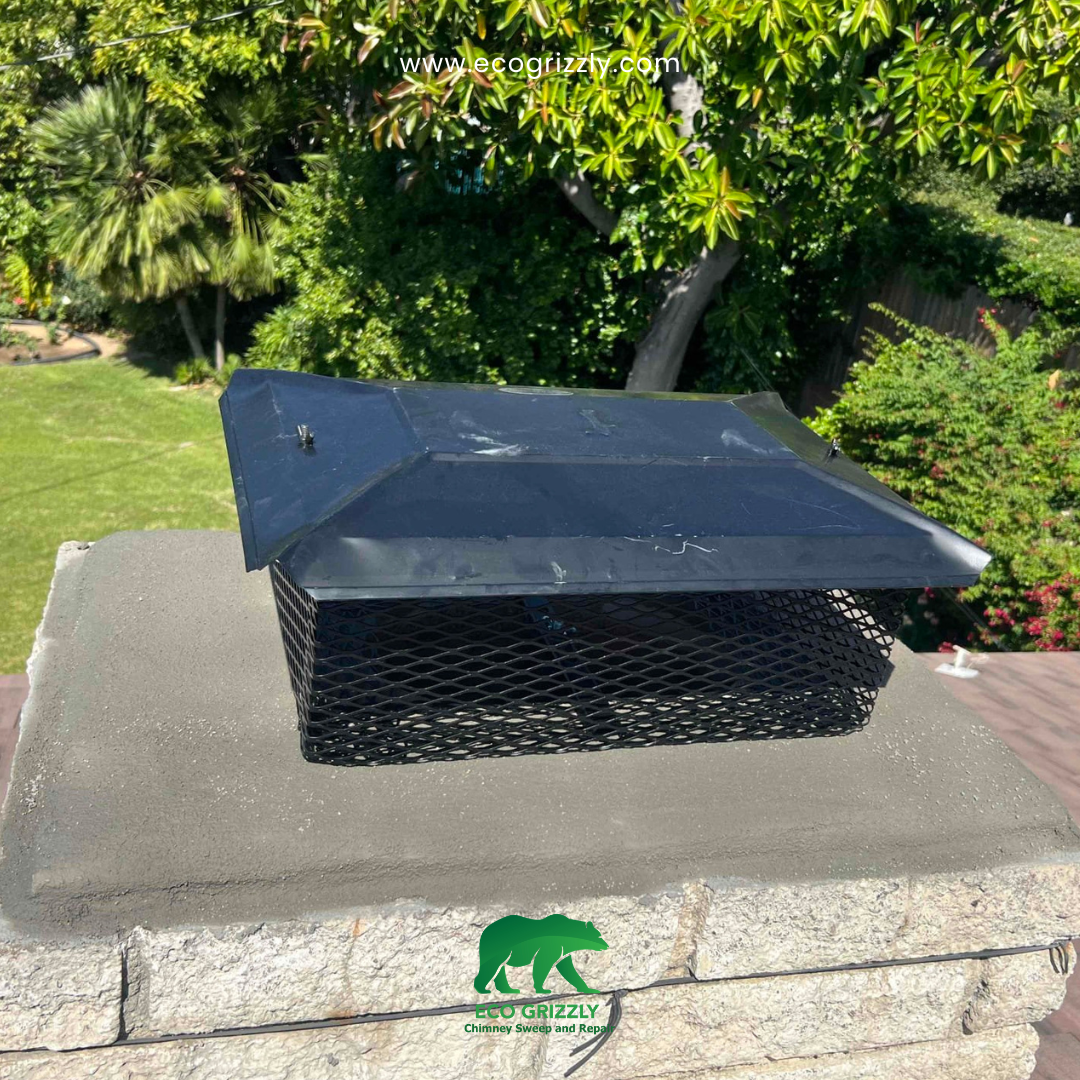 Black metal chimney cap with mesh sides on a stone and concrete chimney with green trees