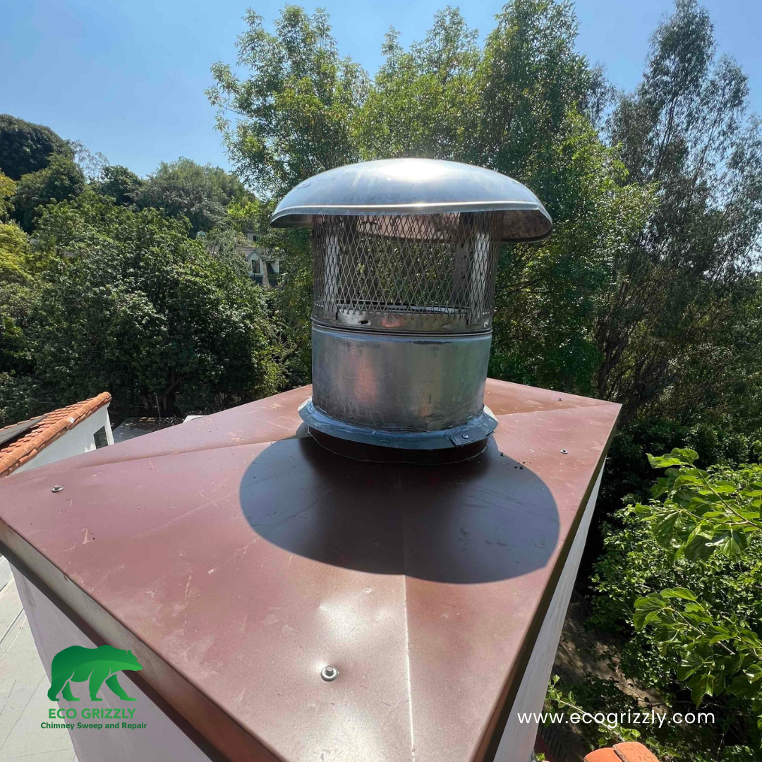 Metal chimney cap with mesh sides on a copper-colored roof surrounded by green trees
