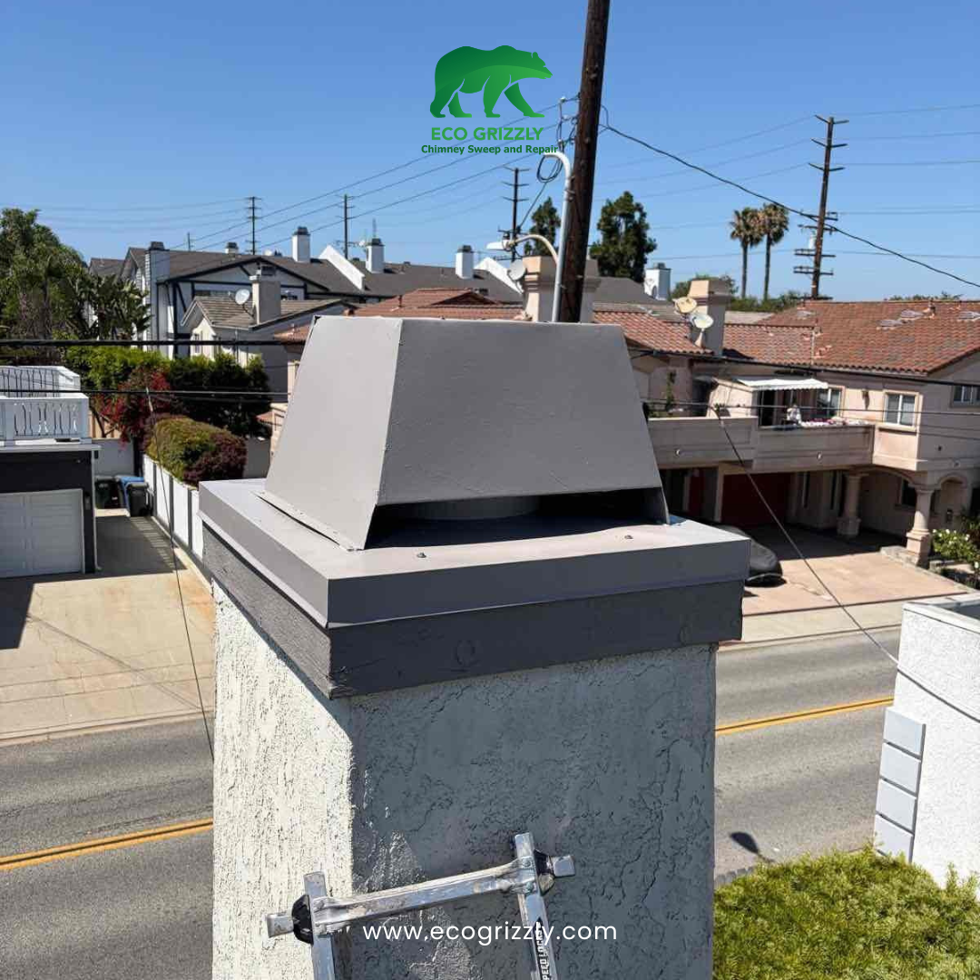 Gray angular chimney cap installed on a stucco chimney with ladder leaning against it