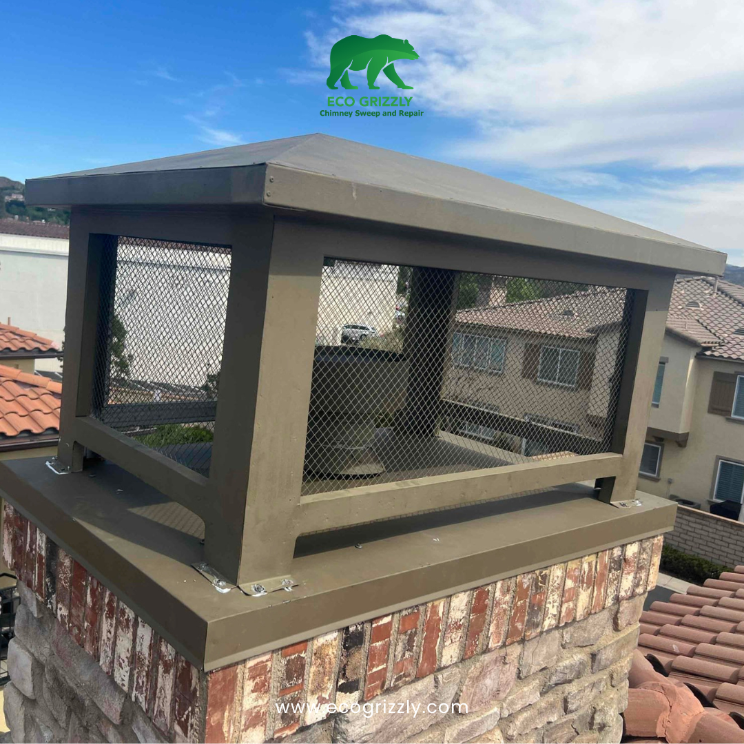 Dark brown metal chimney cap with mesh sides on a red brick chimney with tiled roofs behind