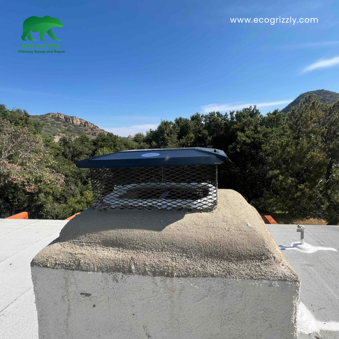 Black metal chimney cap on a stucco chimney with rolling hills and blue sky behind